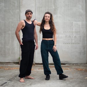 May include: Two people wearing casual clothing stand in front of a concrete wall. One person wears a black tank top and black pants. The other person wears a black crop top and teal pants. The word "RAYA" is visible on the wall.