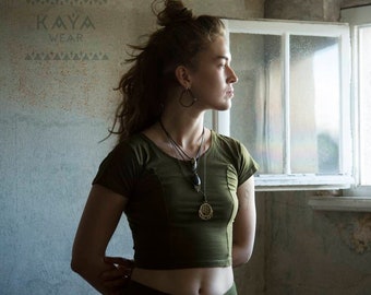 May include: A woman wearing a green cropped t-shirt with a long necklace and a pendant. She is standing in a room with a window behind her. The text "KAYA WEAR" is visible on the wall.