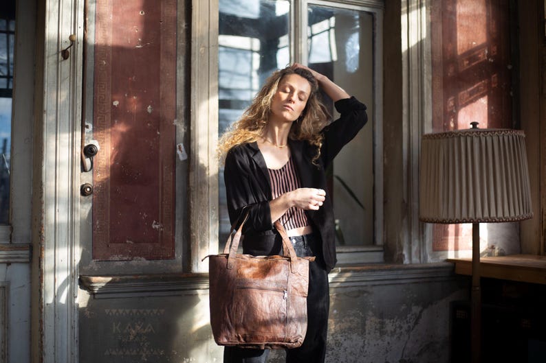 Puede incluir: Un bolso tote de cuero marr&oacute;n con un bolsillo con cremallera, llevado por una persona con una blazer negra y una camiseta a rayas. El bolso tiene un aspecto desgastado y texturizado. El fondo incluye una puerta y una ventana vintage.