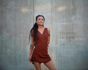 May include: A woman wearing a rust-colored wrap dress with a V-neckline and sleeveless design. She has long, dark hair with dreadlocks and is standing in front of a gray concrete wall. The text "KAYA WEAR" is visible on the wall.