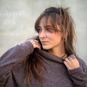 Könnte beinhalten: Eine Frau trägt einen dunkelvioletten Pullover mit hohem Ausschnitt und strukturiertem Strick. Sie trägt mehrere Silberringe und einen Nasenring. Das Logo "KAYA WEAR" ist oben links im Bild zu sehen.