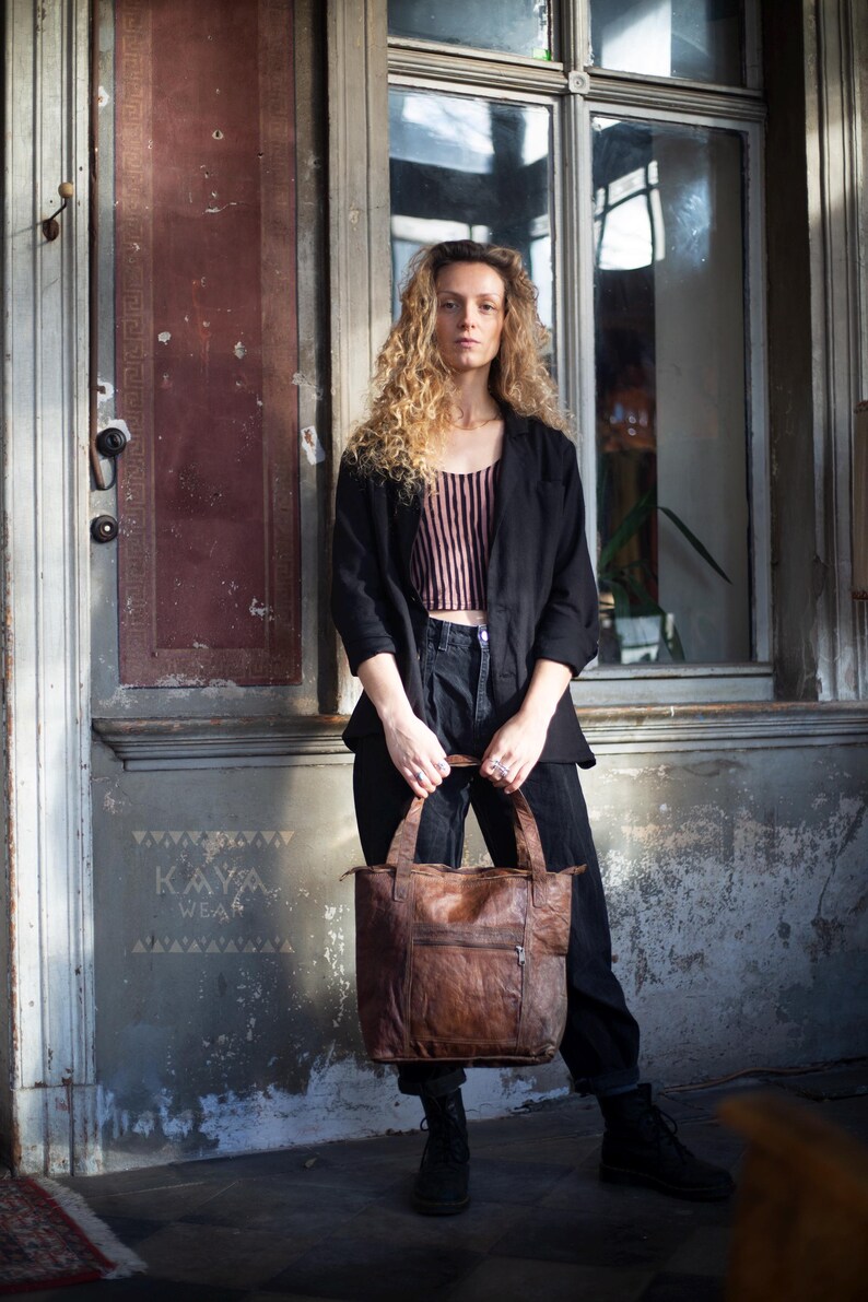Puede incluir: Un bolso tote de cuero marr&oacute;n con un bolsillo con cremallera se sostiene frente a una persona. El bolso tiene un aspecto desgastado y texturizado. La persona lleva una blazer negra, pantalones negros y una camiseta a rayas. La palabra "KAYA WEA" es visible en la pared.
