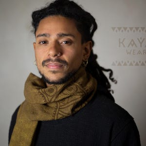 May include: A person with dreadlocks is wearing a brown scarf with a geometric pattern. The scarf is wrapped around their neck and is visible against a black shirt. The text "KAYA WEAR" is visible in the background.