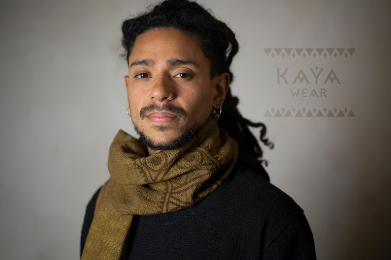 May include: A person with dreadlocks is wearing a brown scarf with a geometric pattern. The scarf is wrapped around their neck and is visible against a black shirt. The text "KAYA WEAR" is visible in the background.