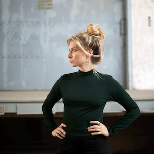Puede incluir: Una mujer con el pelo rubio recogido en un moño lleva una camiseta de cuello alto verde oscuro. Está de pie frente a una pared gris con el texto "KAYA WEAR" escrito en ella.