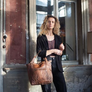 Puede incluir: Un bolso tote de cuero marr&oacute;n con una superficie texturizada. El bolso tiene un asa superior y un bolsillo frontal. La persona de la imagen lleva una blazer negra, una camiseta a rayas y pantalones negros.