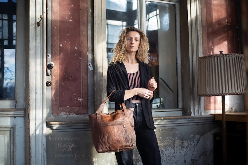 Puede incluir: Un bolso tote de cuero marr&oacute;n con una superficie texturizada. El bolso tiene un asa superior y un bolsillo frontal. La persona de la imagen lleva una blazer negra, una camiseta a rayas y pantalones negros.