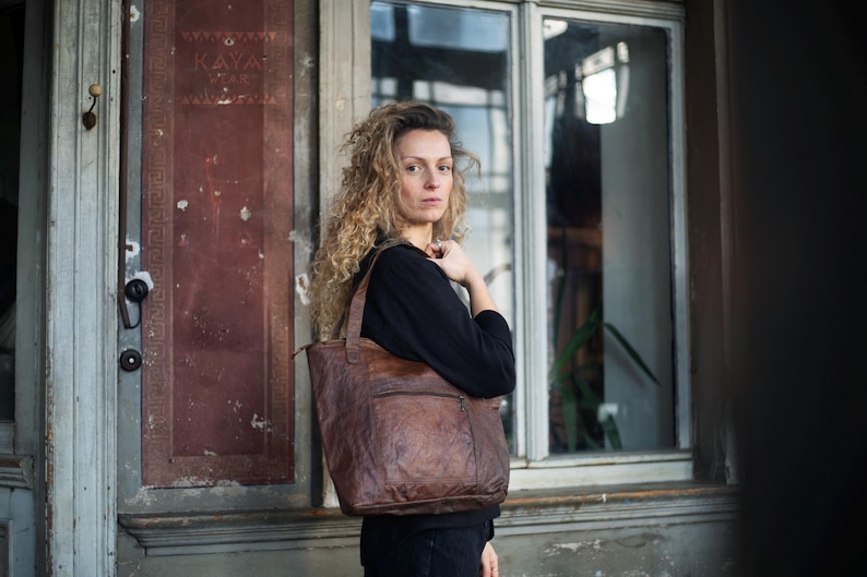 Puede incluir: Un bolso tote de cuero marr&oacute;n con superficie texturizada se lleva sobre el hombro. El bolso tiene forma rectangular con un bolsillo delantero y cierre de cremallera. La mujer lleva un top negro.