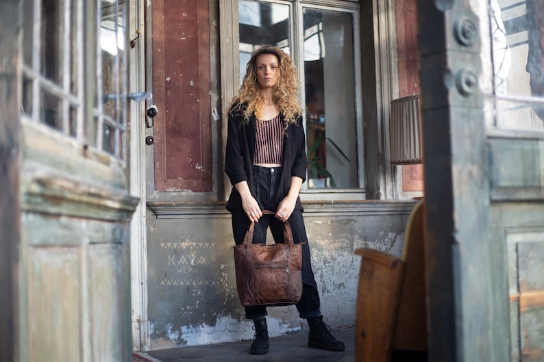 Puede incluir: Una mujer sostiene un bolso tote de cuero marr&oacute;n con un bolsillo con cremallera. Lleva una blazer negra, pantalones negros y botas negras. El bolso tiene un aspecto desgastado. El fondo incluye una puerta y una ventana desgastadas.
