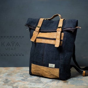 May include: A black backpack with tan leather accents. The backpack has adjustable straps, a top handle, and a roll-top closure. The front features a tan leather pocket and straps with buckles. The brand "KAYA WEAR" is visible.