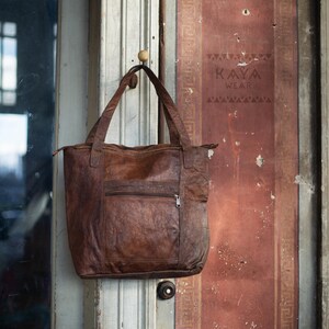 Puede incluir: Un bolso tote de cuero marr&oacute;n con un bolsillo frontal con cremallera y asas dobles, colgado de una puerta de madera. El bolso tiene un aspecto texturizado y un cierre de cremallera. La puerta tiene el texto "KAYA WEAR" impreso.