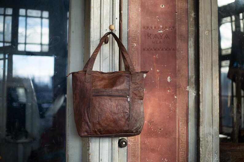 Puede incluir: Un bolso tote de cuero marr&oacute;n con un bolsillo frontal con cremallera y asas dobles, colgado de una puerta de madera. El bolso tiene un aspecto texturizado y un cierre de cremallera. La puerta tiene el texto "KAYA WEAR" impreso.