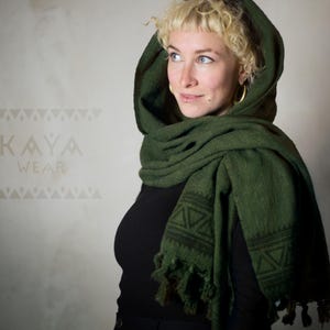 Pode incluir: Uma mulher usando um cachecol com capuz verde escuro com um padrão geométrico. O cachecol é feito de um tecido macio e tecido e tem uma borda franjada. O cachecol é usado sobre uma camisa preta.