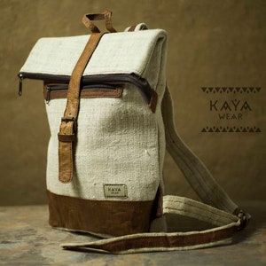 May include: A beige canvas backpack with brown leather straps and accents. The backpack has a flap closure and a zippered pocket on the front. The brand name "KAYA WEAR" is printed on a tag attached to the backpack.