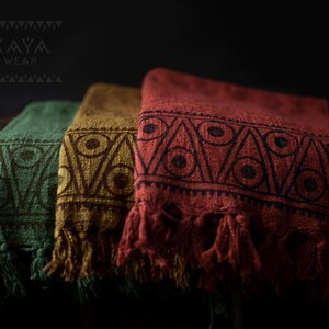 May include: Three woven cotton throws with fringe, featuring a geometric pattern in shades of green, brown, and red. The throws are stacked on a wooden surface. The text "KAYA WEAR" is visible in the top left corner of the image.