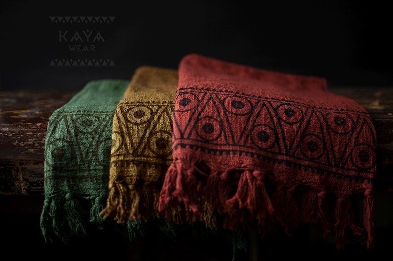 May include: Three woven cotton throws with fringe, featuring a geometric pattern in shades of green, brown, and red. The throws are stacked on a wooden surface. The text "KAYA WEAR" is visible in the top left corner of the image.