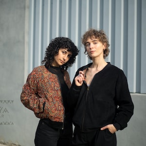 Puede incluir: Dos personas con chaquetas bomber. Una chaqueta tiene un estampado de cachemira en tonos rojos, naranjas y marrones. La otra chaqueta es de color negro liso. Las chaquetas están abrochadas con cremallera y tienen un corte relajado. La marca "KAYA WEAR" es visible.