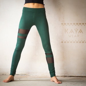 May include: Green leggings with mesh panels. The leggings have a high waist and a slim fit. The mesh panels are located on the sides of the legs. The text "KAYA WEAR" is printed on the wall in a tan color.