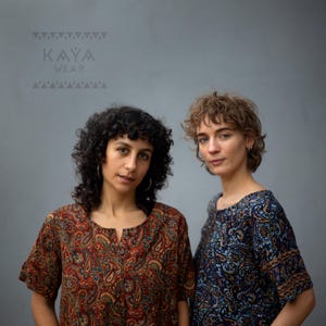 May include: Two people wearing paisley print tops. One top is red and brown, the other is blue and brown. The tops have short sleeves and a scoop neckline. The brand name "KAYA WEAR" is printed in grey at the top of the image.