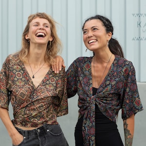May include: Two women wearing colourful patterned wrap tops. The tops are tied at the waist and have a V-neckline. The women are smiling and looking at the camera. The text "KAYA WEAR" is visible in the top right corner of the image.