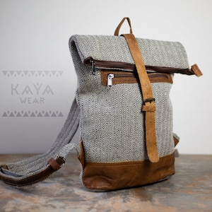 May include: A gray and white herringbone fabric backpack with brown leather accents and a zippered flap closure. The backpack has a brown leather strap with a buckle. The brand name "KAYA WEAR" is printed on the front of the bag.
