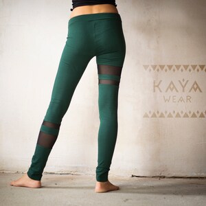 May include: Green leggings with sheer mesh panels. The leggings feature a high waist and a slim fit. The brand name "KAYA WEAR" is visible in a tan color on the wall behind the leggings. The leggings are made of a soft, stretchy material.