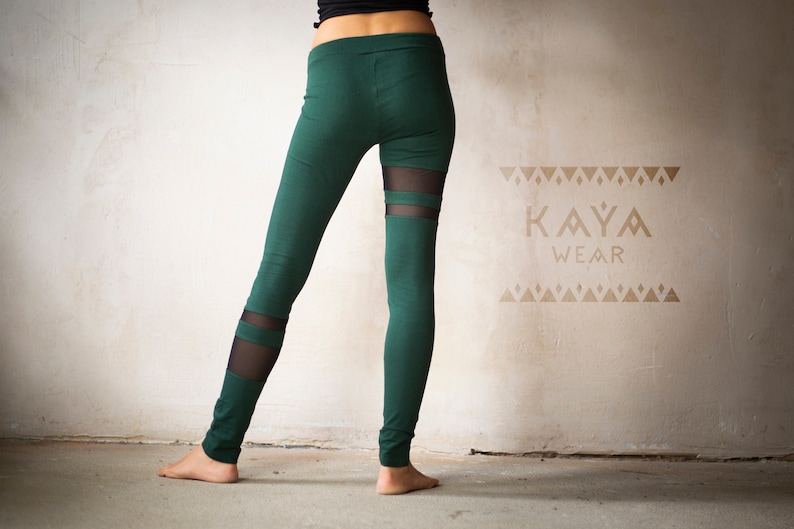 May include: Green leggings with sheer mesh panels. The leggings feature a high waist and a slim fit. The brand name "KAYA WEAR" is visible in a tan color on the wall behind the leggings. The leggings are made of a soft, stretchy material.