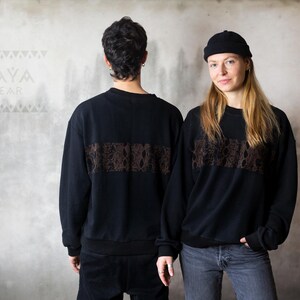 May include: Two people wearing black sweatshirts with a brown patterned band across the chest. The text "KAYA WEAR" is visible on the wall behind them.