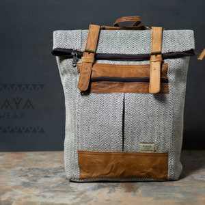 May include: A gray and white herringbone patterned backpack with brown leather accents and straps. The backpack has a zippered closure and a small leather patch with the text "KAYA WEAR" on the front.