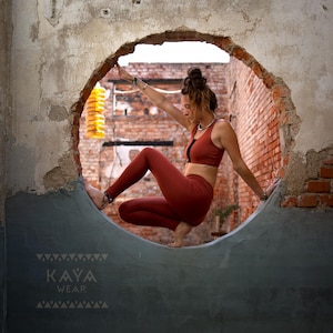 Peut inclure: Une personne vêtue d'un soutien-gorge de sport et d'un legging couleur rouille pose dans une ouverture circulaire dans un mur de briques usé. Le mur est partiellement brisé, révélant la maçonnerie et un aperçu de l'extérieur. La marque "KAYA WEAR" est visible.