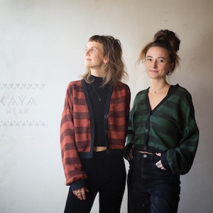 Può includere: Due donne indossano cardigan a righe. Il cardigan a sinistra è rosso e nero, e il cardigan a destra è verde e nero. Entrambi i cardigan sono corti e hanno un collo a V.