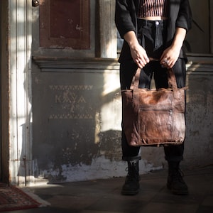 Puede incluir: Un bolso tote grande de cuero marr&oacute;n con aspecto desgastado. El bolso tiene dos asas y un bolsillo frontal. La persona que sostiene el bolso lleva pantalones y botas negras. El fondo es una pared desgastada con la palabra "KAYA" estampada.