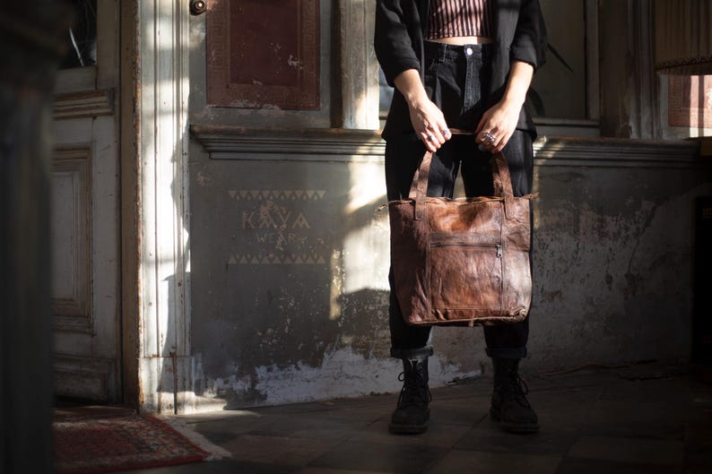 Puede incluir: Un bolso tote grande de cuero marr&oacute;n con aspecto desgastado. El bolso tiene dos asas y un bolsillo frontal. La persona que sostiene el bolso lleva pantalones y botas negras. El fondo es una pared desgastada con la palabra "KAYA" estampada.