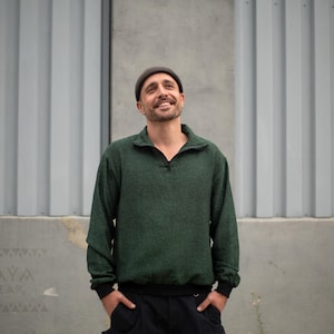 May include: A dark green pullover sweater with a quarter-zip collar and black trim. The sweater is worn with a dark beanie and black pants. The background is a gray industrial wall.