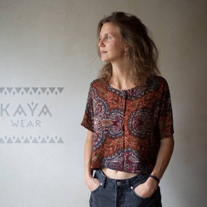 Könnte beinhalten: Eine Frau trägt ein rotes, blaues und goldenes Paisley-Muster-Crop-Top mit Knopfleiste. Das Oberteil ist aus einem leichten Stoff gefertigt und hat eine lockere Passform. Der Text "KAYA WEAR" ist im Hintergrund sichtbar.