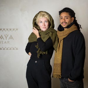 May include: Two people wearing scarves. The woman is wearing a green scarf with a hood and a black top. The man is wearing a brown scarf and a black shirt. The text "KAYA WEAR" is printed on a white wall behind them.