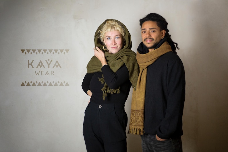 May include: Two people wearing scarves. The woman is wearing a green scarf with a hood and a black top. The man is wearing a brown scarf and a black shirt. The text "KAYA WEAR" is printed on a white wall behind them.