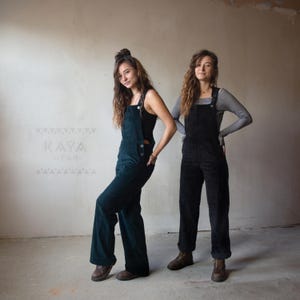 Könnte beinhalten: Zwei Personen tragen Cord-Overalls. Einer ist in Dunkelgrün, der andere in Schwarz. Die Overalls haben verstellbare Träger und Knopfverschlüsse. Der grüne Overall wird mit einem schwarzen Top kombiniert, während der schwarze Overall mit einem gestreiften Langarmshirt getragen wird.