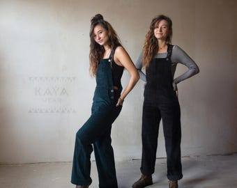 Könnte beinhalten: Zwei Personen tragen Cord-Overalls. Einer ist in Dunkelgrün, der andere in Schwarz. Die Overalls haben verstellbare Träger und Knopfverschlüsse. Der grüne Overall wird mit einem schwarzen Top kombiniert, während der schwarze Overall mit einem gestreiften Langarmshirt getragen wird.