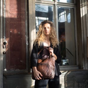 Puede incluir: Una persona sosteniendo un bolso tote de cuero marr&oacute;n con un dise&ntilde;o acolchado. El bolso tiene un bolsillo frontal y dos asas. La persona lleva una chaqueta negra y una camiseta a rayas. El fondo incluye una ventana y una puerta.