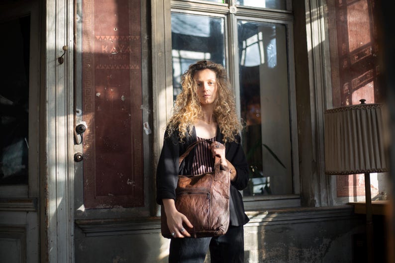 Puede incluir: Una persona sosteniendo un bolso tote de cuero marr&oacute;n con un dise&ntilde;o acolchado. El bolso tiene un bolsillo frontal y dos asas. La persona lleva una chaqueta negra y una camiseta a rayas. El fondo incluye una ventana y una puerta.