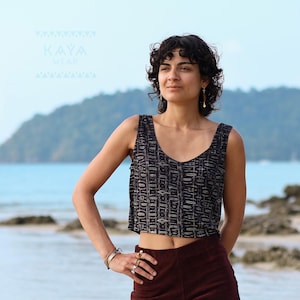May include: A woman wearing a black and white patterned cropped tank top with a burgundy skirt. She is standing on a beach with a blue ocean and green hills in the background.