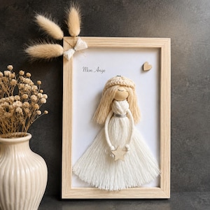 Peut inclure: Une décoration murale encadrée représentant une poupée ange en macramé avec des cheveux en fil crème et une robe blanche. L'ange tient une petite étoile en bois. Le texte Mon Ange est imprimé sur le fond blanc avec un petit cœur en bois.