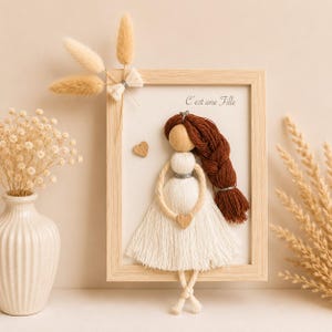 May include: A framed wall art piece featuring a doll made of white yarn and a wooden bead head with long braided brown yarn hair. The doll wears a white yarn dress and holds a small wooden heart. The text C'est une Fille is printed above.