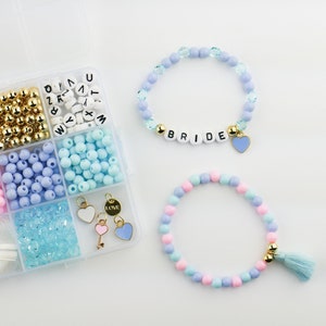 May include: A clear plastic storage box filled with colorful beads, letter beads that spell "BRIDE", and a charm. There are two bracelets made with the beads, one with a blue heart charm and the other with a blue tassel.