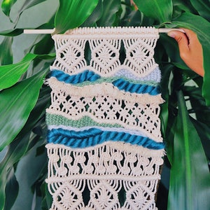 May include: A beige macrame wall hanging with blue and green yarn accents. The hanging features a woven pattern with fringe at the bottom.
