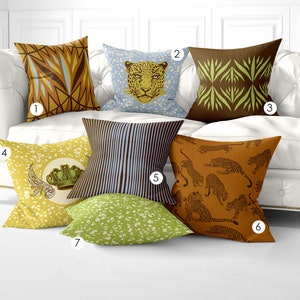May include: Seven decorative throw pillows with various patterns and colors, including brown, yellow, blue, and green. The pillows are arranged on a white couch. The pillows feature geometric patterns, animal prints, and a crown design.