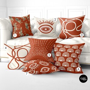 May include: Six decorative throw pillows with various patterns in shades of orange and white. The pillows are arranged on a white couch. The pillows feature designs such as a tiger, an eye, a sunburst, and abstract shapes.