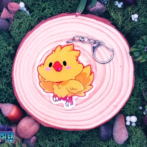 May include: A yellow cartoon chick keyring with a red beak and pink cheeks. The keyring is attached to a silver metal clasp. The chick is on a wooden slice surrounded by green moss and foliage. The keyring is from the "Mookie Monster Shop".