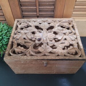 May include: A wooden box with a carved floral design on the lid. The box is light brown in color and has a small knob on the front.